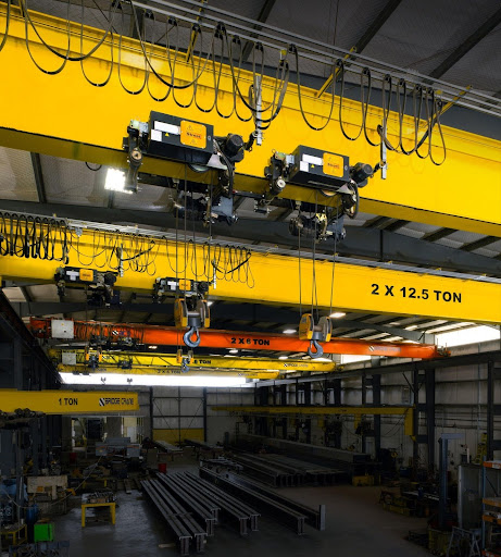 Interior of an industrial facility with several large yellow and orange overhead cranes labeled with their load capacities, above metal beams and machinery that demonstrate how an electric chain hoist works in heavy lifting operations.