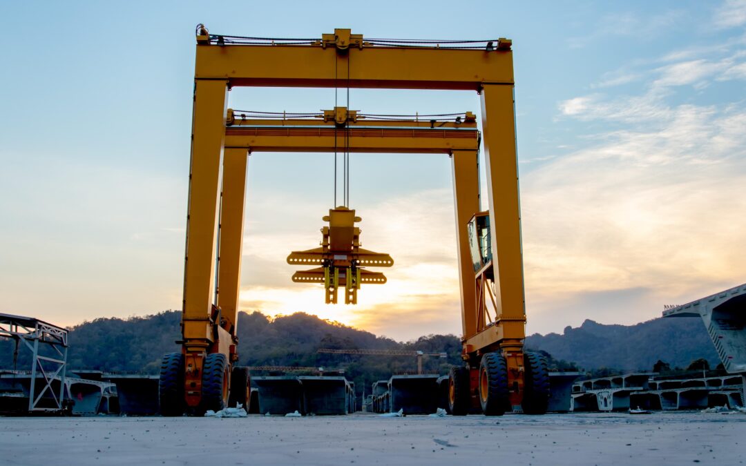 A large yellow gantry crane stands on a concrete surface at sunset, with hills and scattered construction materials in the background.