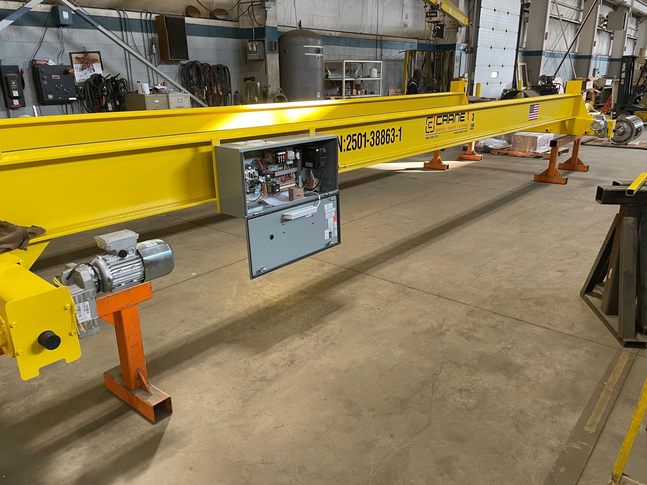 A large yellow overhead crane with an open electrical control panel is positioned indoors on orange stands in an industrial workshop.