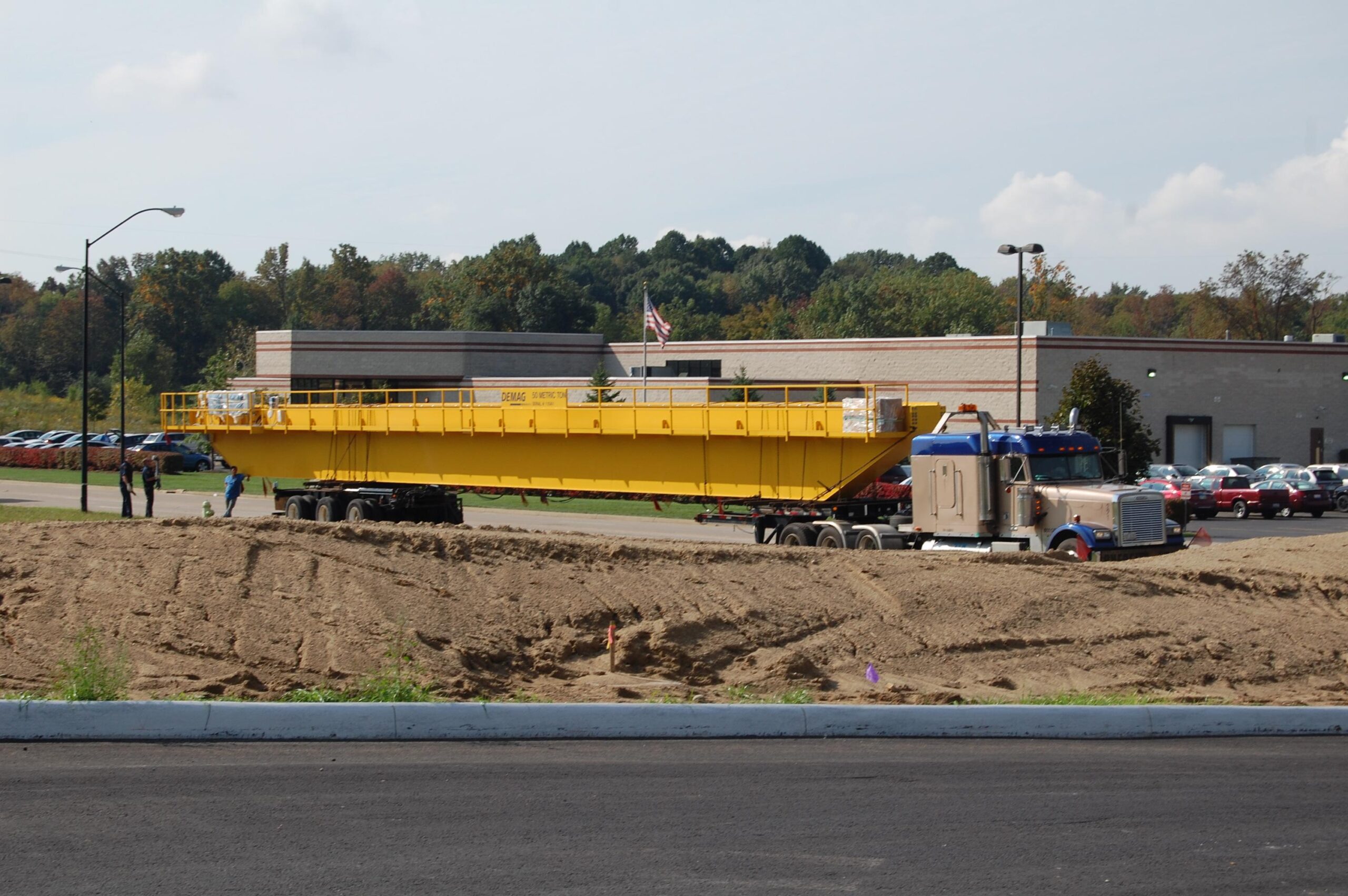 A large yellow steel beam is being transported on a flatbed truck near a construction site with a building and parked cars in the background.