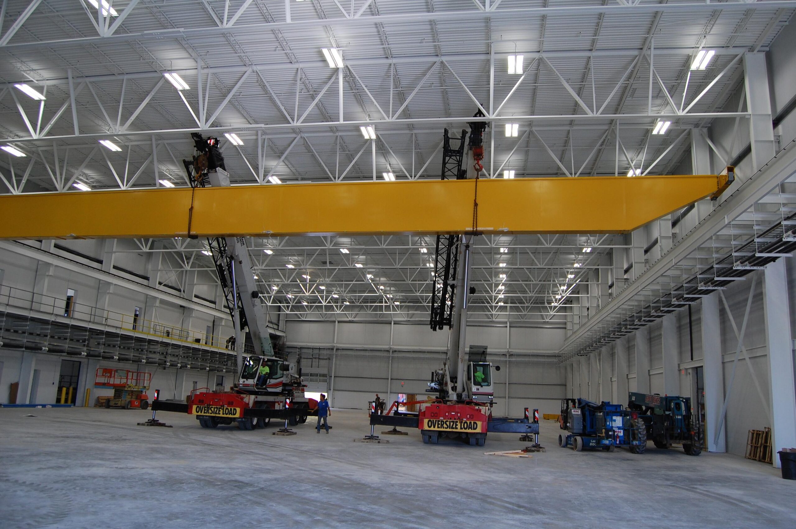 Spacious industrial warehouse featuring new crane solutions, two large construction vehicles, workers, and equipment all under bright overhead lights.