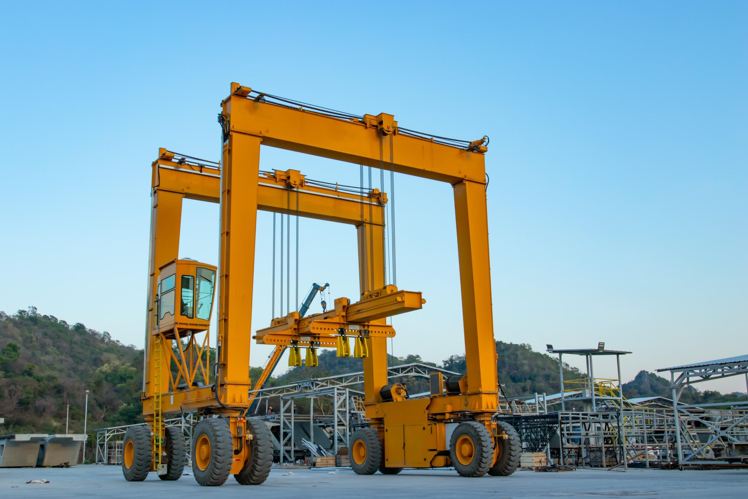 A large yellow gantry crane stands outdoors on a concrete surface, with hills and industrial structures in the background under a clear sky.