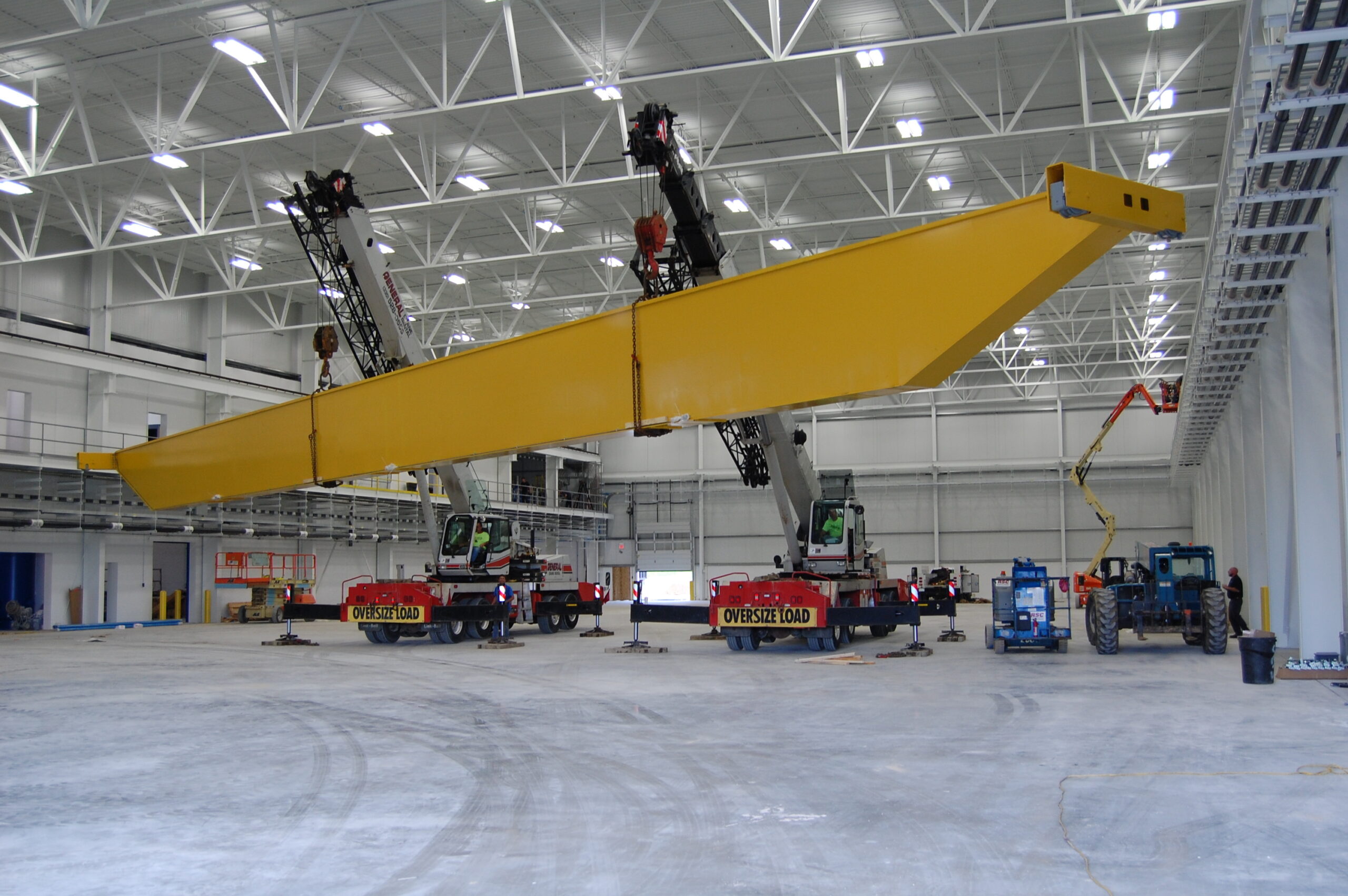 Large yellow steel beam being lifted by two cranes inside a spacious industrial building, with construction vehicles and workers nearby.