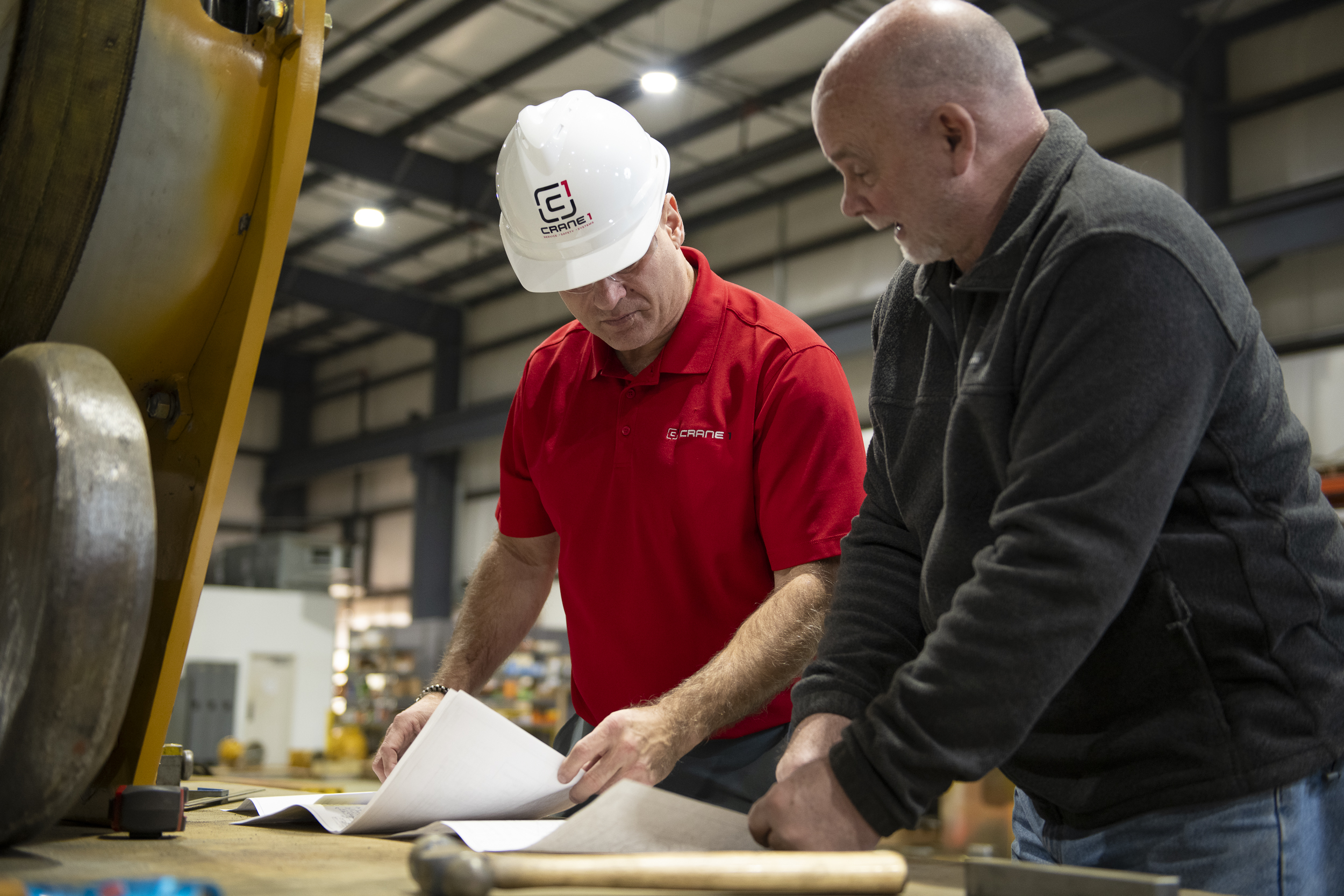 Two men review documents on a workbench inside an industrial facility; one wears a hard hat and red shirt, the other has a dark pullover.