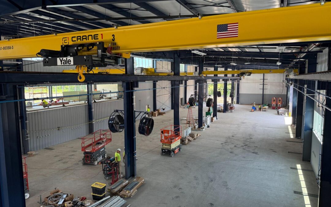 Interior of an industrial warehouse under construction, featuring a yellow overhead crane, construction equipment, tools, and materials scattered on the floor.
