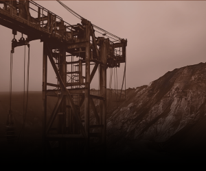 An industrial mining structure with an overhead crane system stands near a rocky hillside under a cloudy sky.