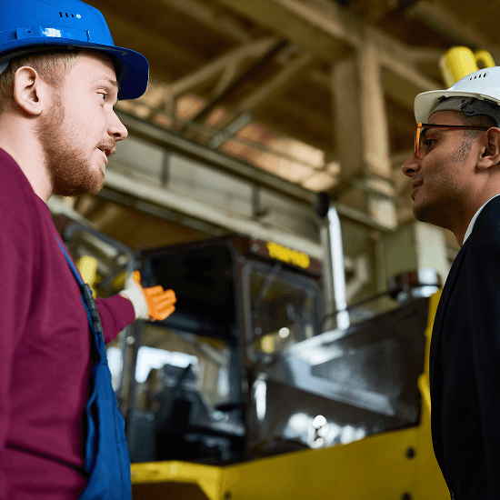 Two men wearing safety helmets and work attire are discussing near industrial machinery inside a factory or warehouse.
