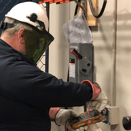 A worker wearing safety gear operates electrical equipment with protective gloves and face shield in an industrial setting.
