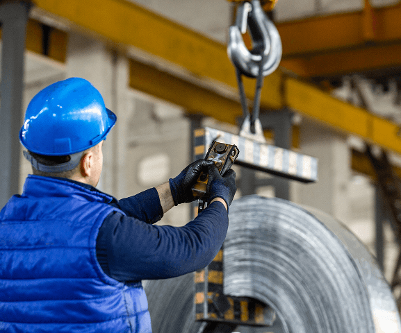 A worker in a blue helmet and vest operates a remote control to lift a large metal coil with an overhead crane inside an industrial facility.