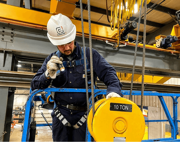 A worker in safety gear inspects a yellow 10-ton hoist on an elevated platform inside an industrial facility.