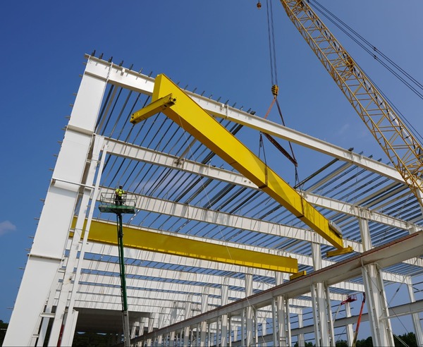 A yellow steel beam is being lifted by a crane onto a white steel frame structure at a construction site under a clear blue sky.