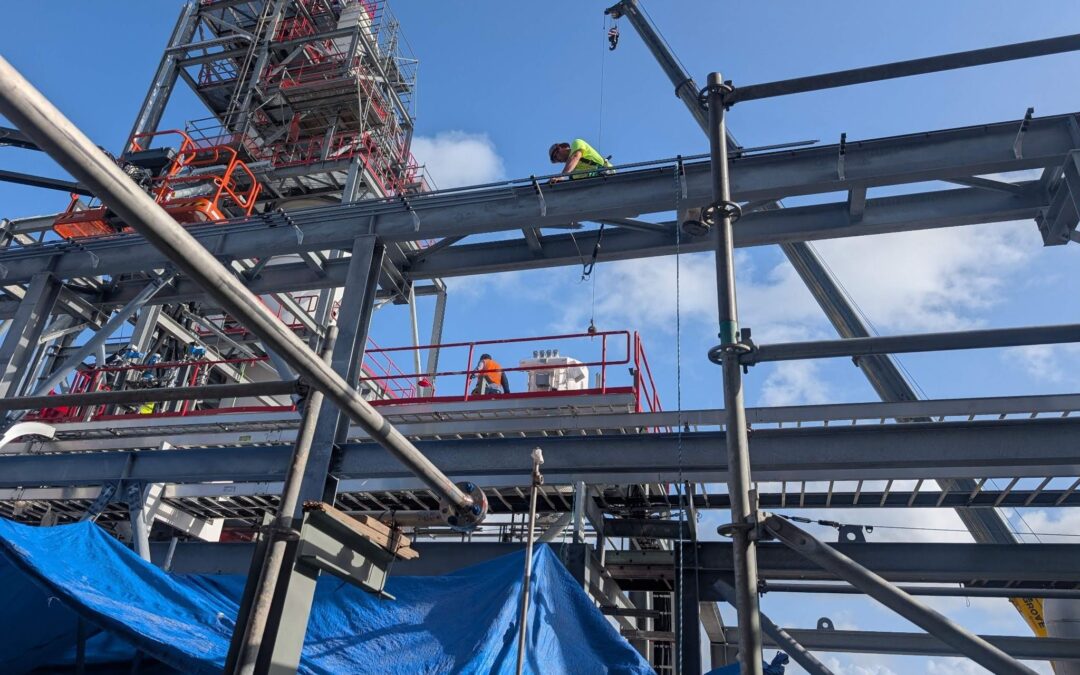 A worker in a safety vest stands on a steel beam at an industrial construction site with scaffolding and blue tarps under a partly cloudy sky.