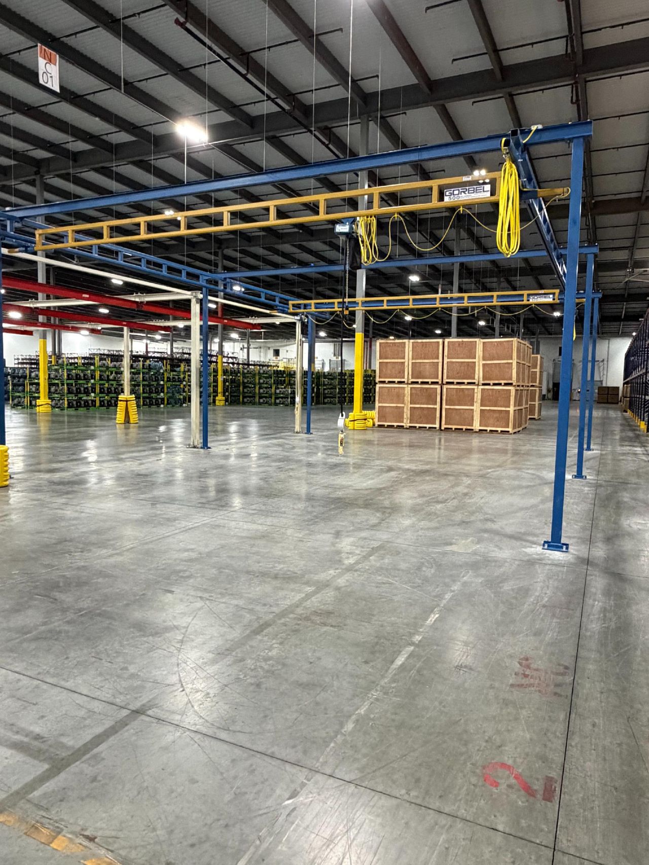 Large warehouse interior with concrete floor, metal beams, yellow cables, stacked wooden crates, and rows of green shelving units in the background.
