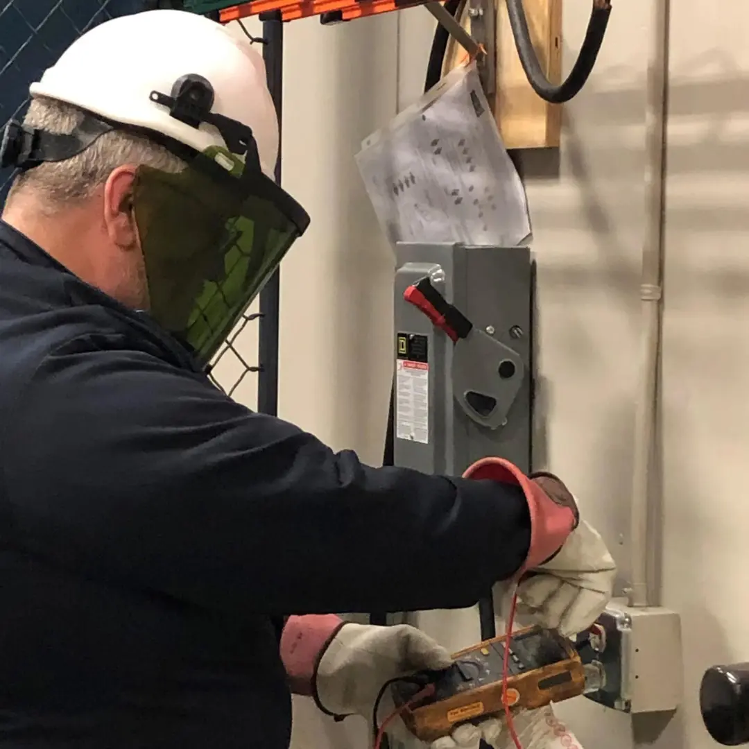 A person wearing a hard hat and face shield uses a multimeter to test an electrical disconnect switch.